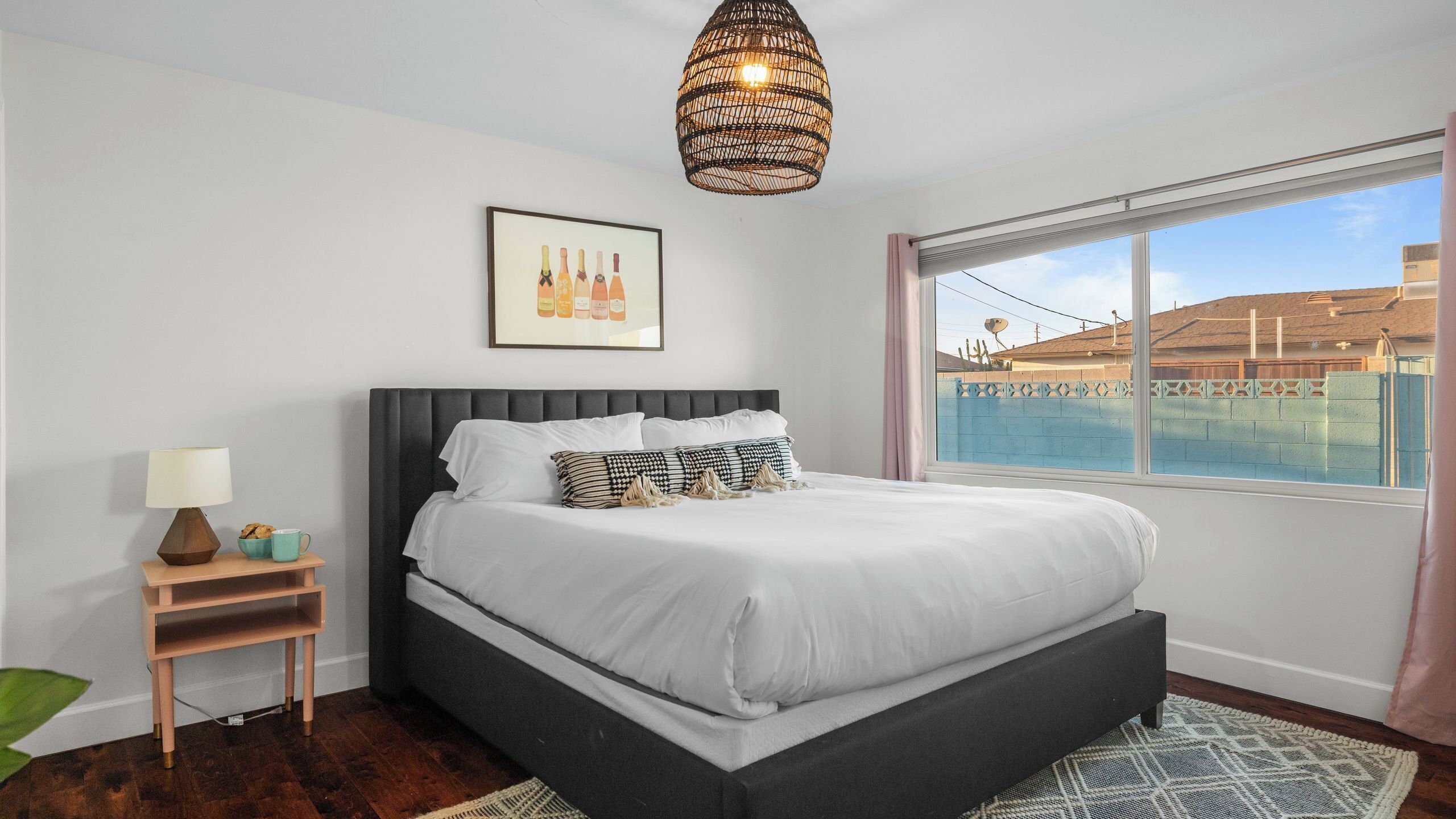 Quarto com uma cama grande, mesa de cabeceira com abajur, quadro emoldurado na parede e vista para um edifício pela janela.