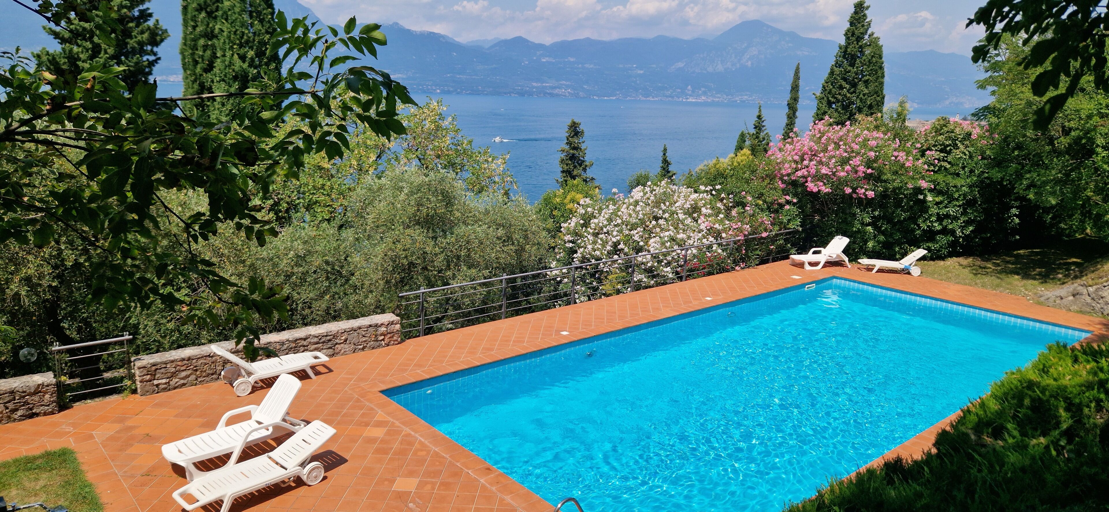 A rectangular swimming pool with a clear blue water, surrounded by a tiled deck. There are two white lounge chairs on the deck. The pool is set against a backdrop of lush greenery and a mountainous landscape.