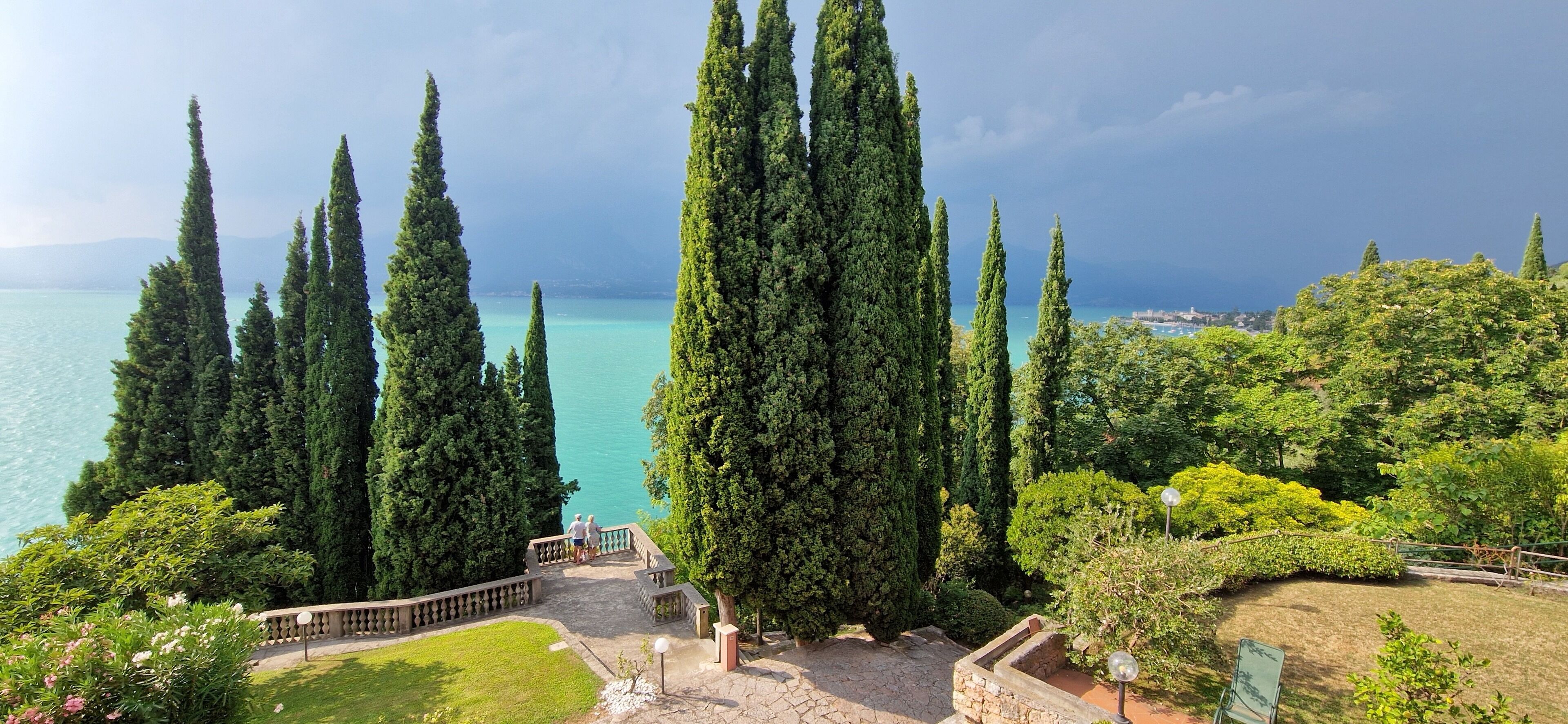 A landscaped garden with tall cypress trees, a stone pathway, and a view of a body of water.