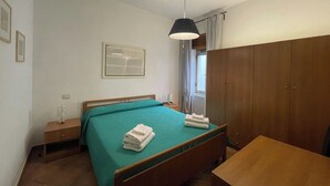 Un dormitorio con cama, mesita de noche, escritorio, armario, lámpara colgante y ventana con cortinas.