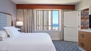 Een hotelkamer met een groot bed, een nachtkastje, een dressoir, een televisie en een raam met uitzicht op een hoog gebouw en de oceaan.