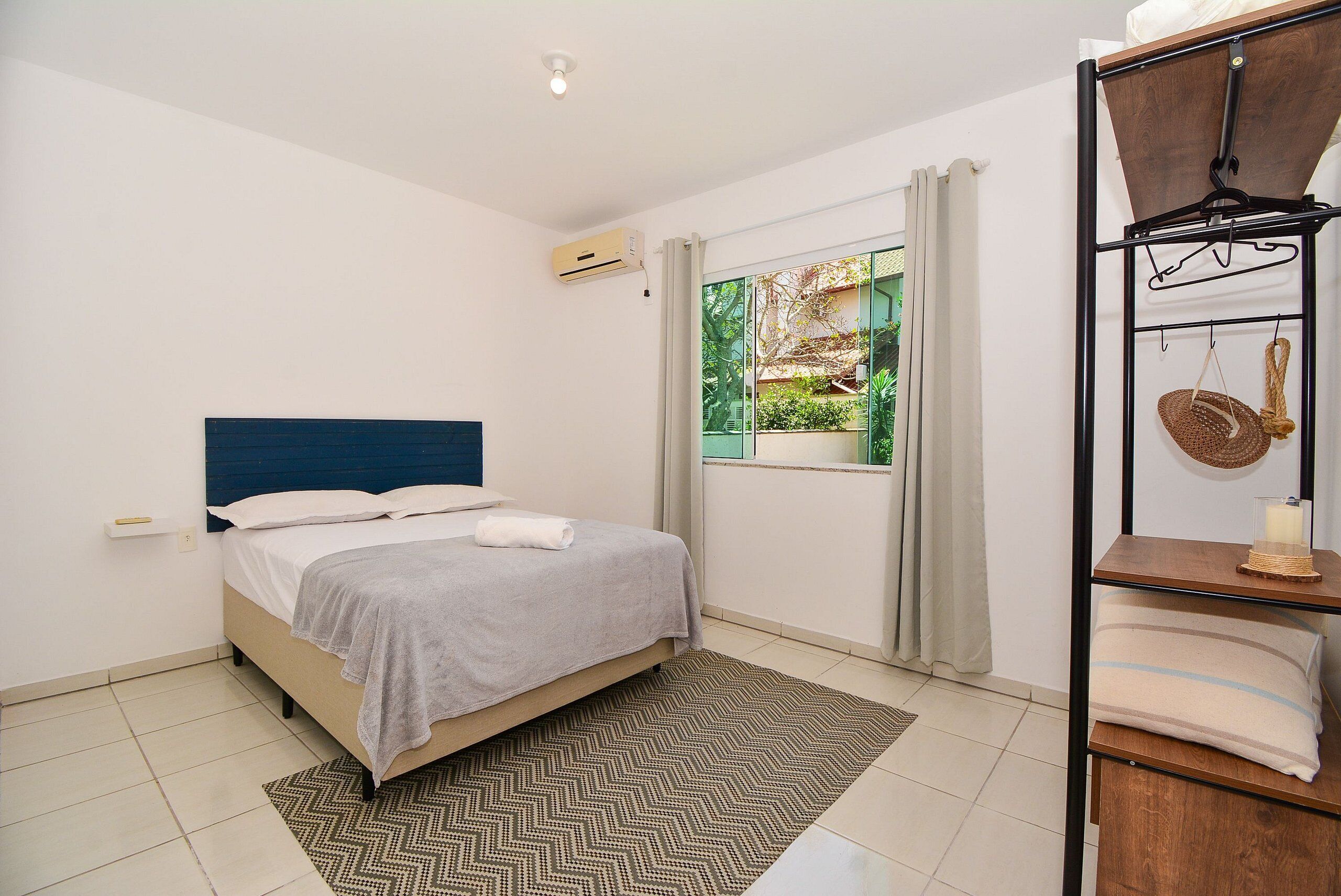 A bedroom with a bed, a window with curtains, a wall-mounted air conditioner, and a shelf with towels.