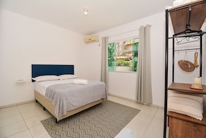 Un dormitorio con cama, ventana con cortinas, aire acondicionado de pared y estante con toallas.