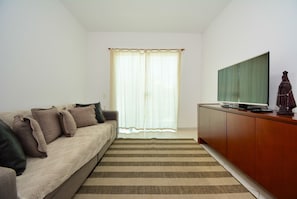 Una sala de estar con un sofá, un televisor sobre un mueble de madera y un ventanal con cortinas traslúcidas.