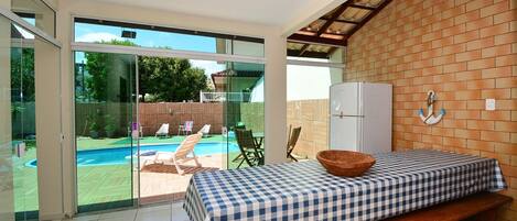 Une pièce avec une table à carreaux, un banc en bois, un réfrigérateur et une vue sur une piscine, visible à travers des portes vitrées.