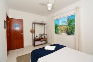 A bedroom with a bed, a wooden wardrobe, a shelf with towels, a ceiling fan, and a window with a view of palm trees. - Casa com piscina a 250m da Praia PLH006 (Palhoça)