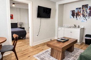 A modern living room with a wooden coffee table, a TV mounted on the wall, and a kitchen area with white cabinets. - The Belmont at PWC (Hot Springs)