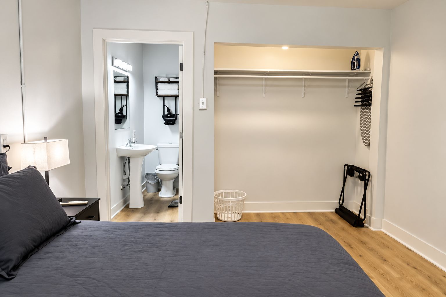 Un dormitorio con cama, mesita de noche, lámpara y baño con lavamanos, inodoro y espejo.