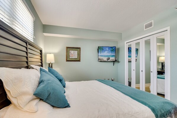 Une chambre à coucher comprenant un lit, une table de chevet, une fenêtre avec des stores, un tableau encadré et une armoire avec un miroir.
