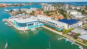 Un puerto deportivo con barcos amarrados, un edificio grande con techo azul y edificios residenciales y comerciales alrededor.
