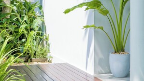 Un patio moderno al aire libre con una terraza de madera, plantas en macetas y una pared blanca con una puerta y luces fijadas a la pared.