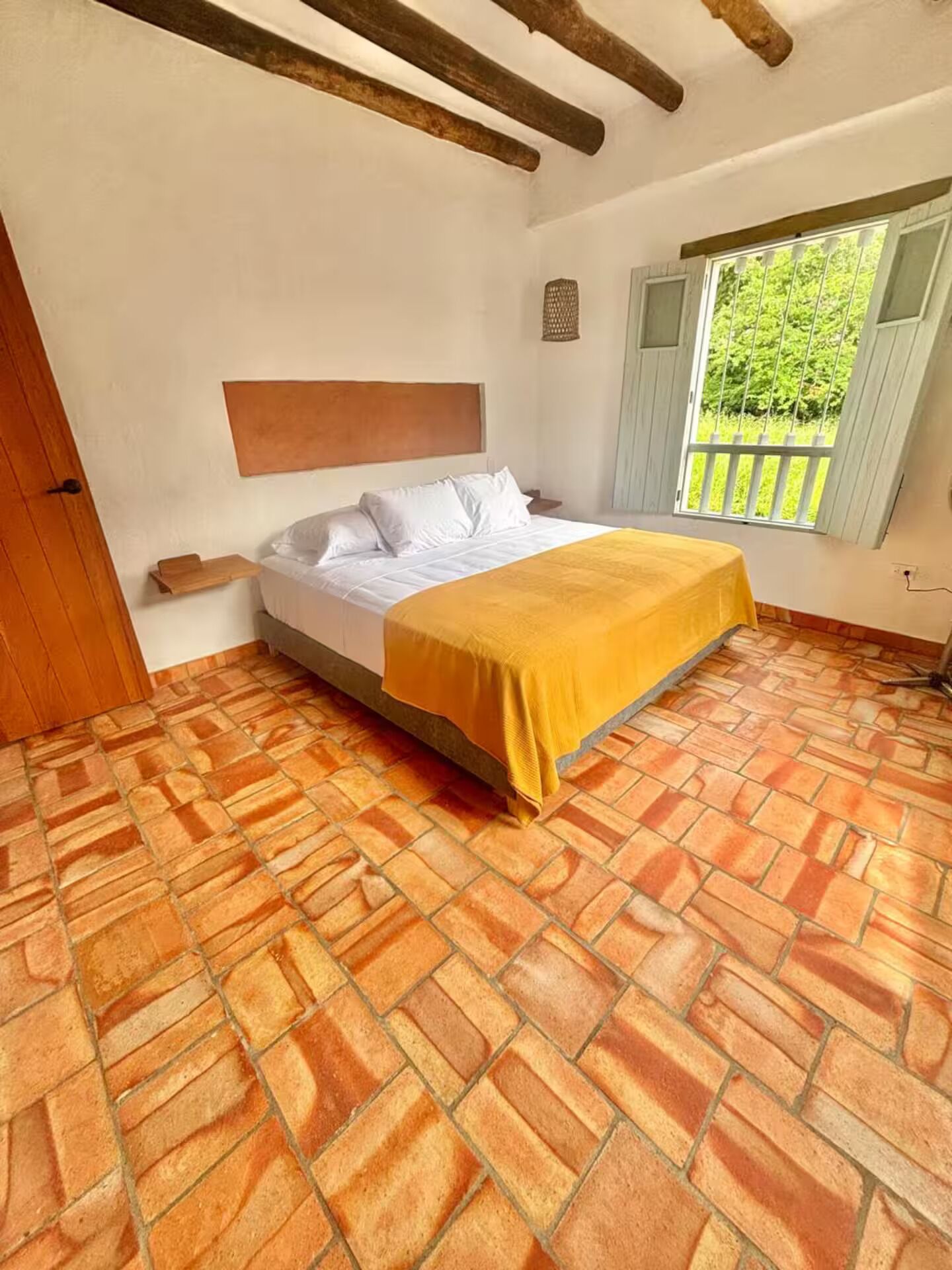 A bedroom with a bed, a wooden headboard, a window with a view of greenery, and a tiled floor.