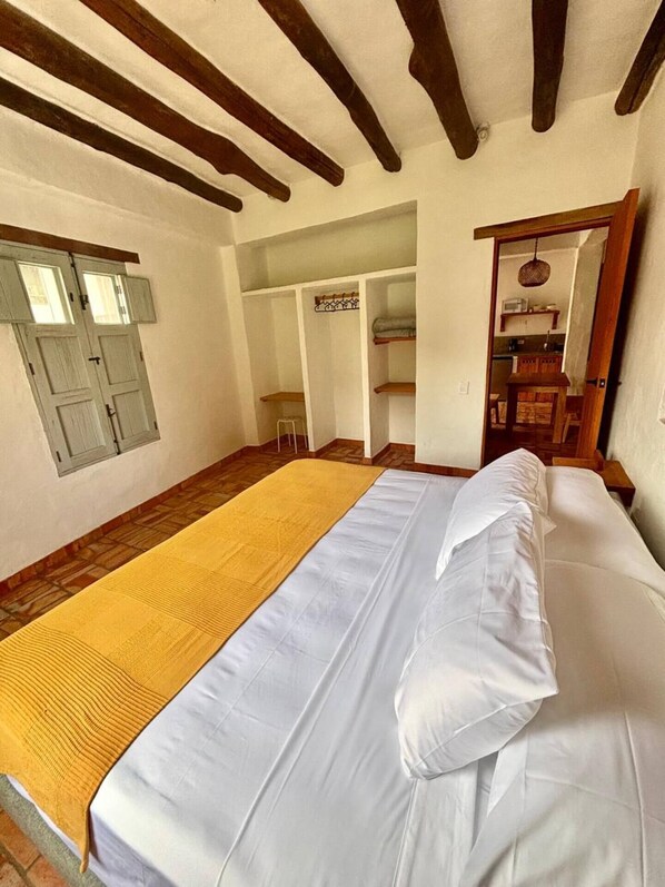 Un dormitorio con cama, una manta amarilla, vigas de madera en el techo y una pequeña kitchenette al fondo.