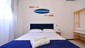 A neatly made bed with white linens and a blue accent, a small bedside table with a plant, and a wooden bunk bed to the right. - Refúgio do Mar Centrinho Ingleses-NIN017 (Florianópolis)