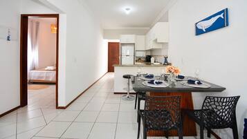 Una cocina moderna con armarios blancos, un comedor con una mesa dispuesta para cuatro comensales, y un dormitorio visible a través de una puerta abierta.