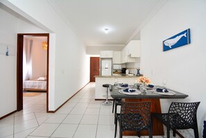 Une cuisine moderne avec des meubles blancs, un coin repas avec une table dressée pour quatre personnes, et une chambre à coucher visible à travers une porte ouverte.