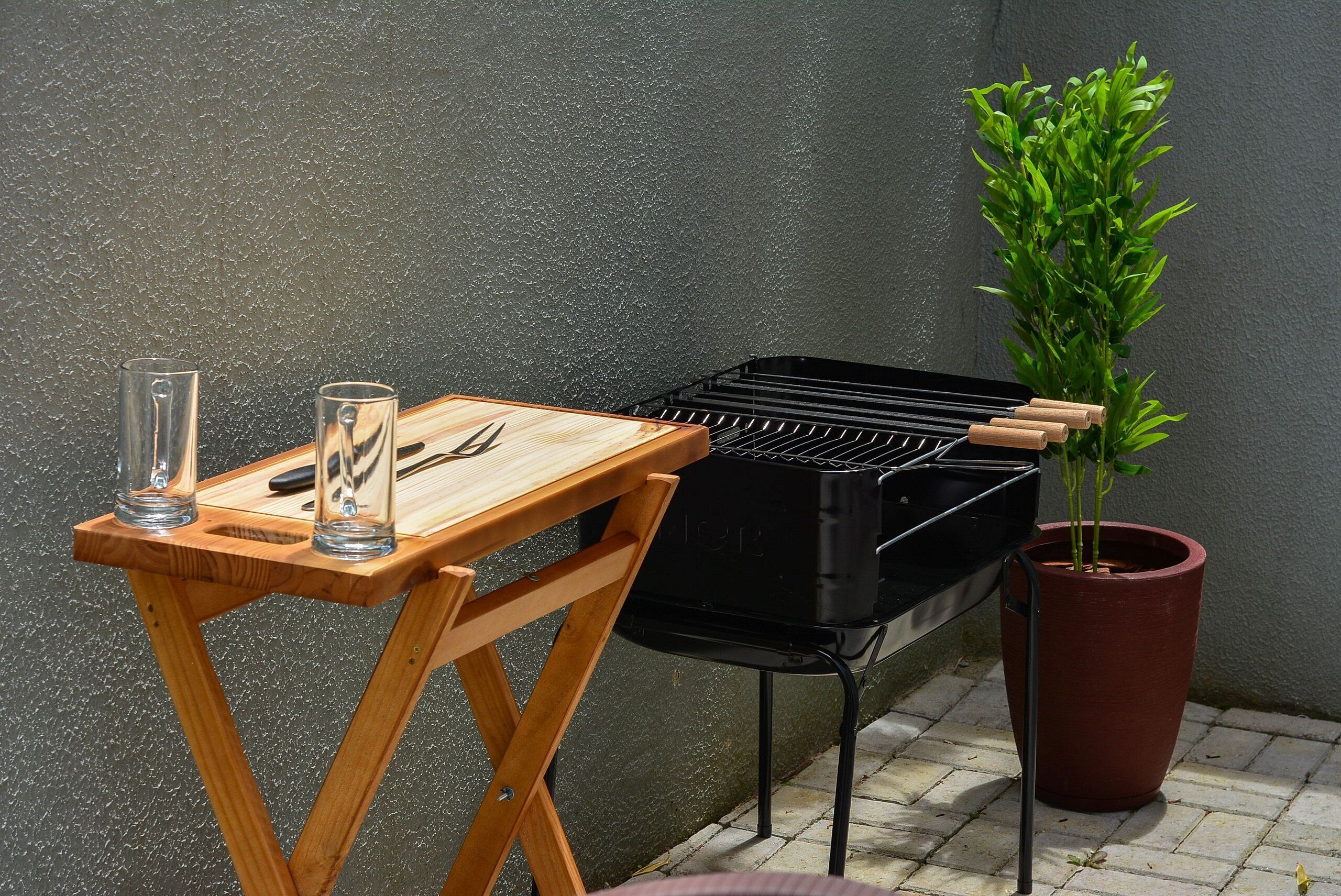 En portabel grill placerad på ett träbord, tillsammans med glas och redskap, bredvid en krukväxt.