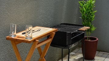 En portabel grill placerad på ett träbord, tillsammans med glas och redskap, bredvid en krukväxt.