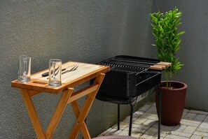Un barbecue portable installé sur une table en bois, avec des verres et des ustensiles à proximité, à côté d’une plante en pot.