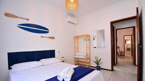 Un lit bien fait, avec du linge de lit blanc et un élément décoratif bleu, une étagère en bois avec des serviettes et un climatiseur fixé au mur.
