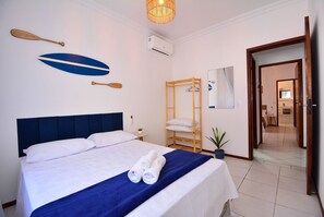 Un lit bien fait, avec du linge de lit blanc et un élément décoratif bleu, une étagère en bois avec des serviettes et un climatiseur fixé au mur.