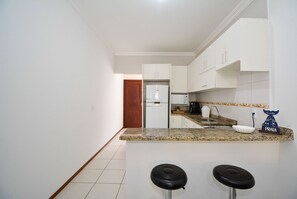 A kitchen with white cabinets, a granite countertop, a refrigerator, a microwave, and a coffee maker. There are two black stools in front of the counter. - Refúgio do Mar Centrinho Ingleses-NIN017 (Florianópolis)