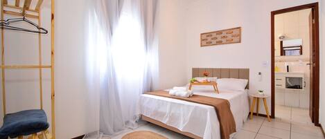 A bedroom with a bed, a wooden hanger rack, a small table, and a bathroom visible through an open door.