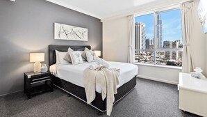 A modern hotel room with a large bed, a city view, a painting on the wall, and a nightstand with a lamp. - Chevron Renaissance - 2 Bed 1 bath Low Floor - 1098 - 2 night - Chevron 2 Bedroom 1 Bathroom Apartment (Surfers Paradise)