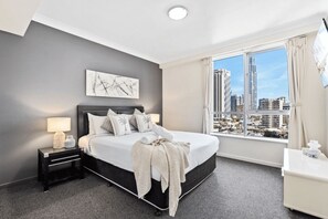 A modern hotel room with a large bed, a city view, a painting on the wall, and a nightstand with a lamp. - Chevron Renaissance - 2 Bed 1 bath Low Floor - 1098 - 3 night - Chevron 2 Bedroom 1 Bathroom Apartment (Surfers Paradise)