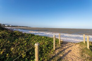 Uma trilha costeira que leva à praia, com vista ampla do oceano e edifícios ao longe.