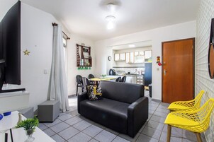 Uma sala de estar com sofá preto, televisão, uma mesinha com uma planta e uma cozinha ao fundo.