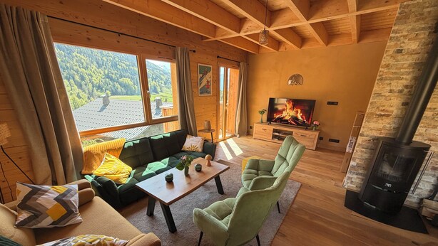 Un salon chaleureux avec un canapé, des fauteuils, une table basse, une cheminée et de grandes fenêtres donnant sur l’extérieur.