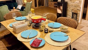 Un coin repas chaleureux, avec une table en bois dressée pour six personnes, entourée de chaises rembourrées. On aperçoit une cheminée au fond, et une affiche décore le mur.