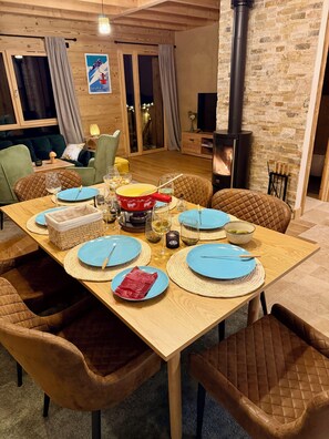 A cozy dining area with a wooden table set for six, surrounded by cushioned chairs. A fireplace is visible in the background, and a poster adorns the wall. - New chalet, absolute calm & mountain views - Near La Clusaz & Grand-Bornand (Saint-Jean-de-Sixt)