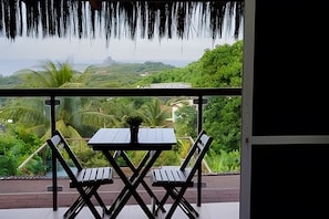 Uma varanda com mesa e cadeiras, oferecendo vista para uma paisagem exuberante, com vegetação e o oceano ao longe.