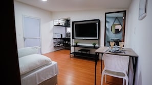 A hotel room with a bed, a TV, a small table with a chair, and a kitchenette area.