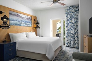 Une chambre d’hôtel avec un lit, deux lampes fixées au mur, un ventilateur de plafond, un tableau, une télévision et un rideau à motifs.