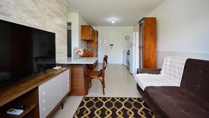 A modern living room with a flat-screen TV, a sofa, a dining area with a wooden table and chairs, and a kitchen with a refrigerator and cabinets. - Apartamento Praia dos Ingleses - NIN015 (Florianópolis)