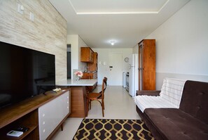A modern living room with a flat-screen TV, a sofa, a dining area with a wooden table and chairs, and a kitchen with a refrigerator and cabinets. - Apartamento Praia dos Ingleses - NIN015 (Florianópolis)