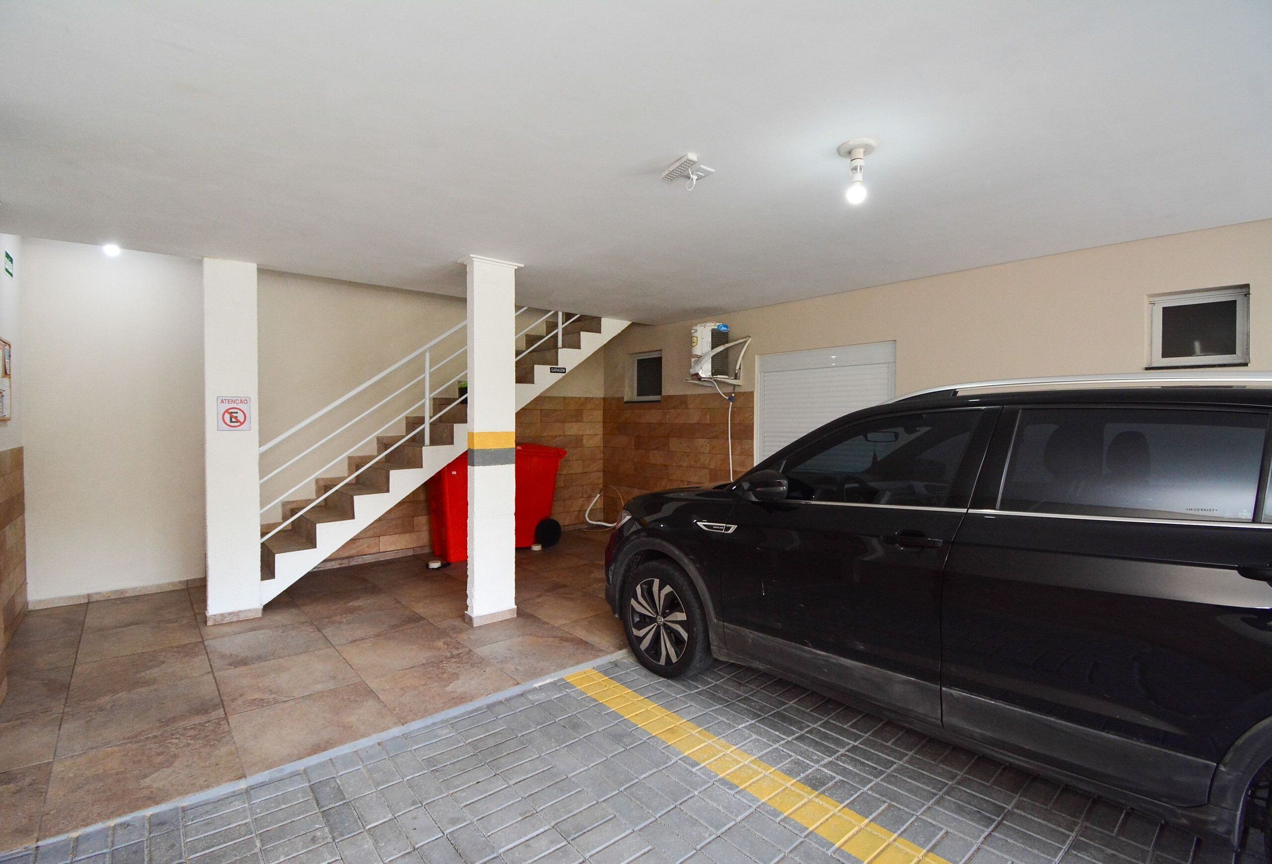 A parking garage with a black SUV, a staircase, and a red trash bin.