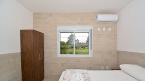 A bedroom with a wooden wardrobe, a bed with a floral bedspread, a window with a view of greenery, and an air conditioning unit. - Apartamento Praia dos Ingleses - NIN015 (Florianópolis)