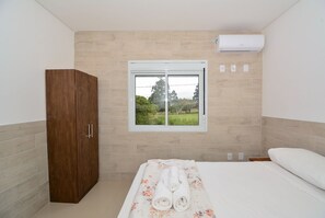 A bedroom with a wooden wardrobe, a bed with a floral bedspread, a window with a view of greenery, and an air conditioning unit. - Apartamento Praia dos Ingleses - NIN015 (Florianópolis)