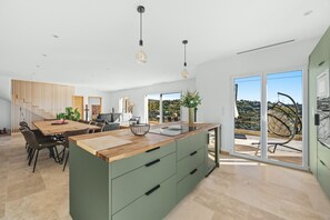 Une cuisine moderne avec un îlot central en bois, un coin repas avec des chaises et une vue sur l’extérieur grâce à de grandes fenêtres.
