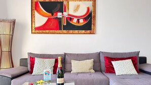 Un salón moderno con un sofá gris, cojines rojos y estampados, una mesa de centro de cristal con fresas y una botella de champán, y un cuadro abstracto grande en la pared.