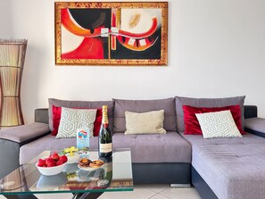 A modern living room with a grey sofa, red and patterned cushions, a glass coffee table with strawberries and a bottle of champagne, and a large abstract painting on the wall. - Luxury Apartment Ocean View by Be Cherish (Albufeira)