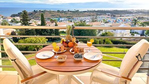 Una mesa redonda de madera con platos de comida, una piña y un vaso de jugo de naranja en un balcón con vistas a un pueblo costero.