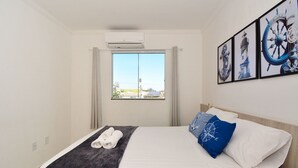 A hotel room with a bed, a window with curtains, a wall-mounted air conditioner, and framed nautical-themed artwork.