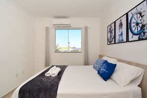 A hotel room with a bed, a window with curtains, a wall-mounted air conditioner, and framed nautical-themed artwork. - Amplo Apartamento Praia Ingleses -NIN013 (Florianópolis)