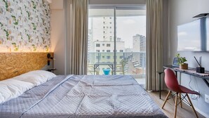 A modern hotel room with a large bed, a desk with a chair, a TV, and a view of the city through large windows. - Apartamento na S c Academia Piscina e Smart Lock (São Paulo)