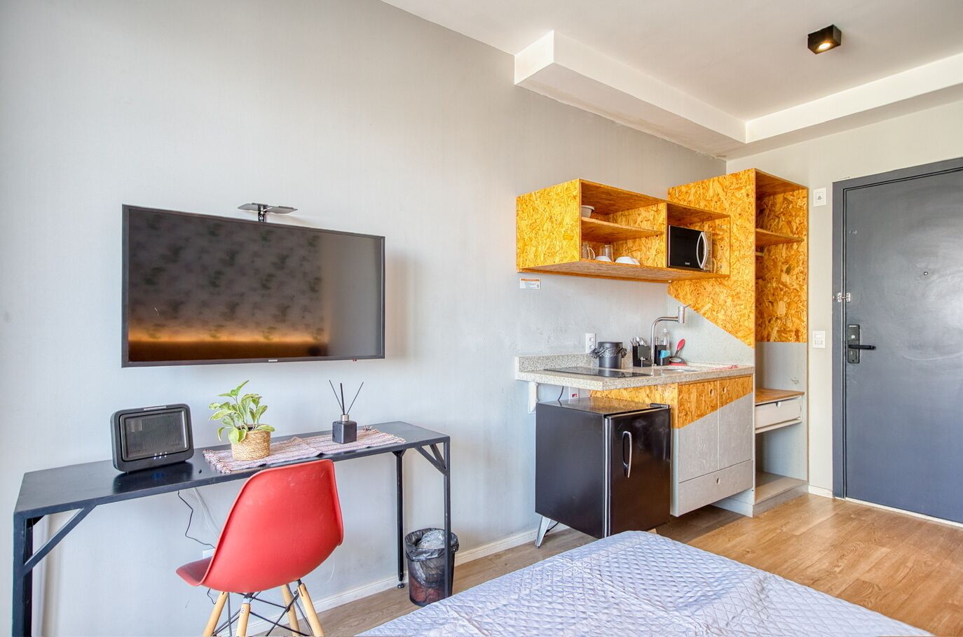 A compact hotel room with a kitchenette, a bed, a desk with a chair, a TV, and a door.